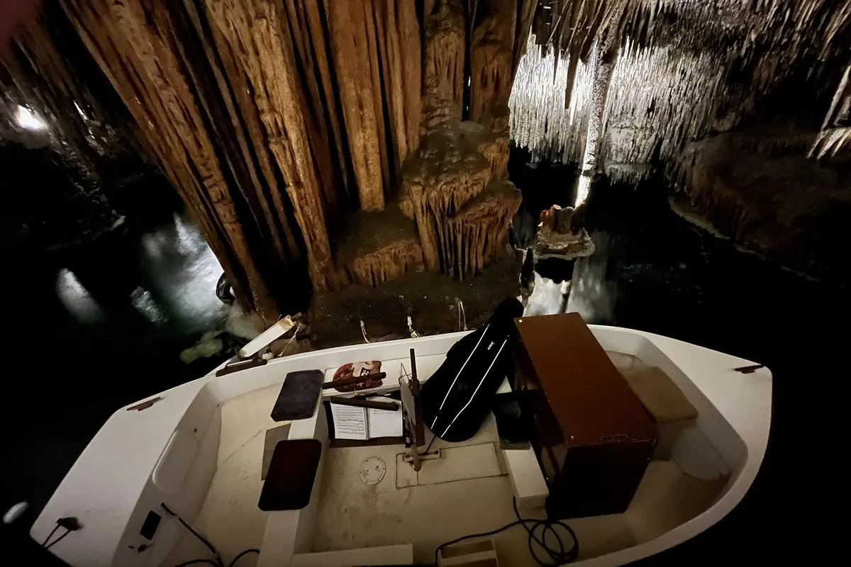 A small white boat on the dark lake in the Drach caves, with a music stand and an instrument case aboard, under a wall of rippled stalactites.