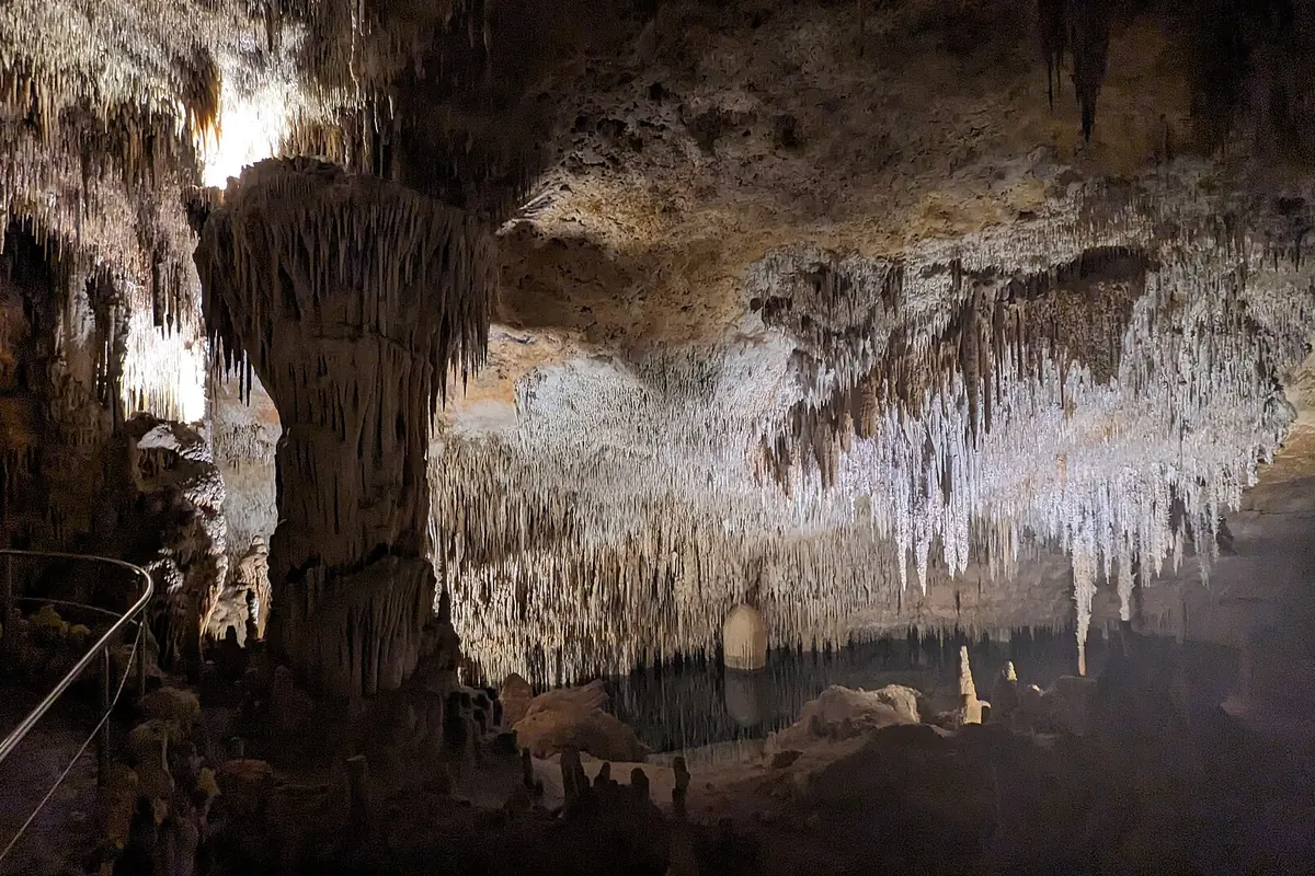 A broad gallery with a railing along the path, a thick column in the middle and fine stalactites above the water.