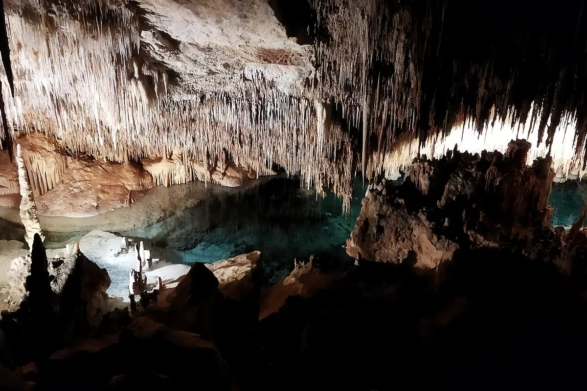 A pool of turquoise lit water at the foot of a stalactite-hung chamber, with rock formations dark around it.
